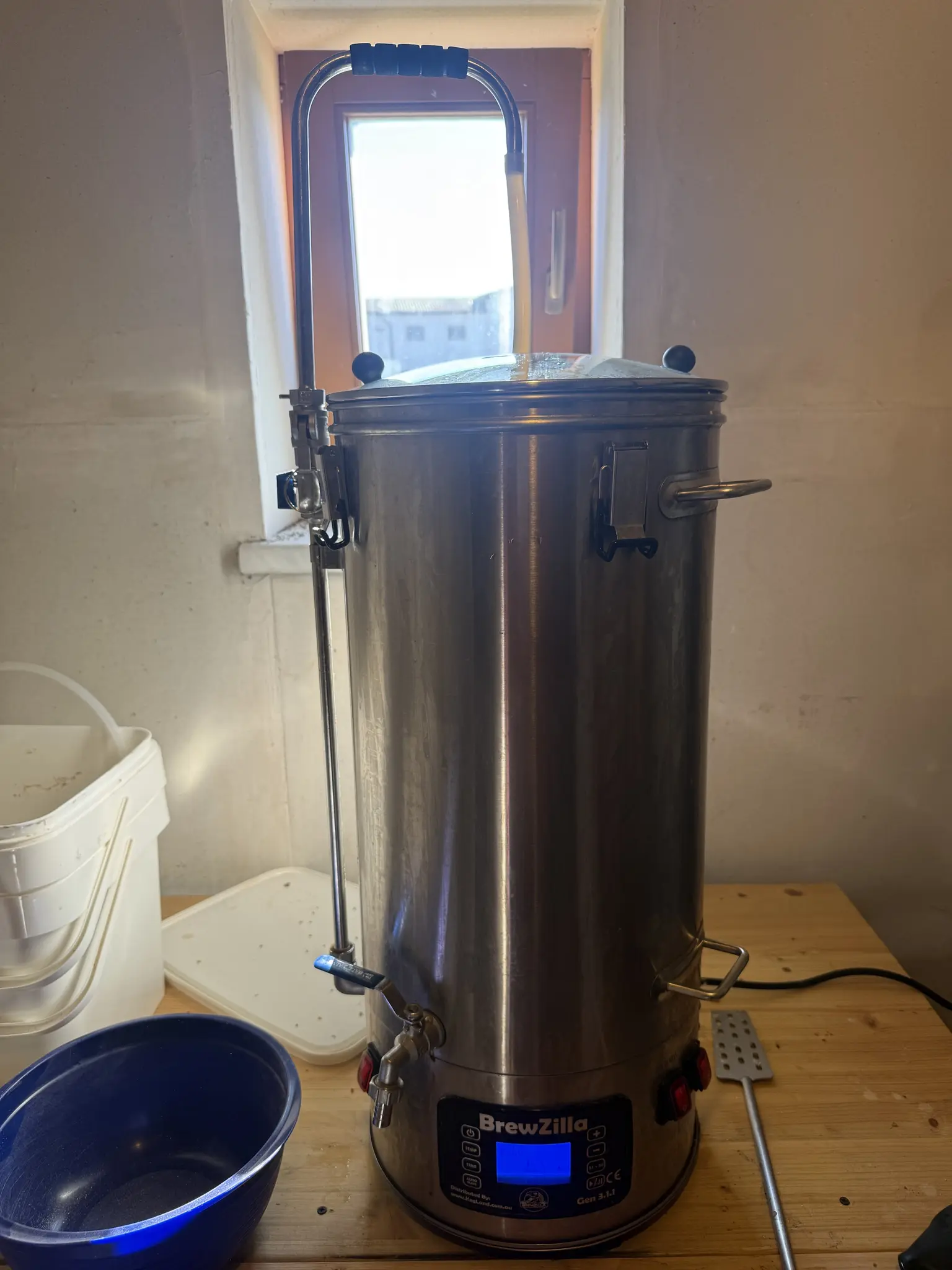 Brewzilla Braukessel auf einem Holtisch, daneben diverse Gefäße und Schläuche und ein Maische-Paddel, dahinter ein kleines Fenster, durch den man den Bauernhof vom Nachbarn sieht