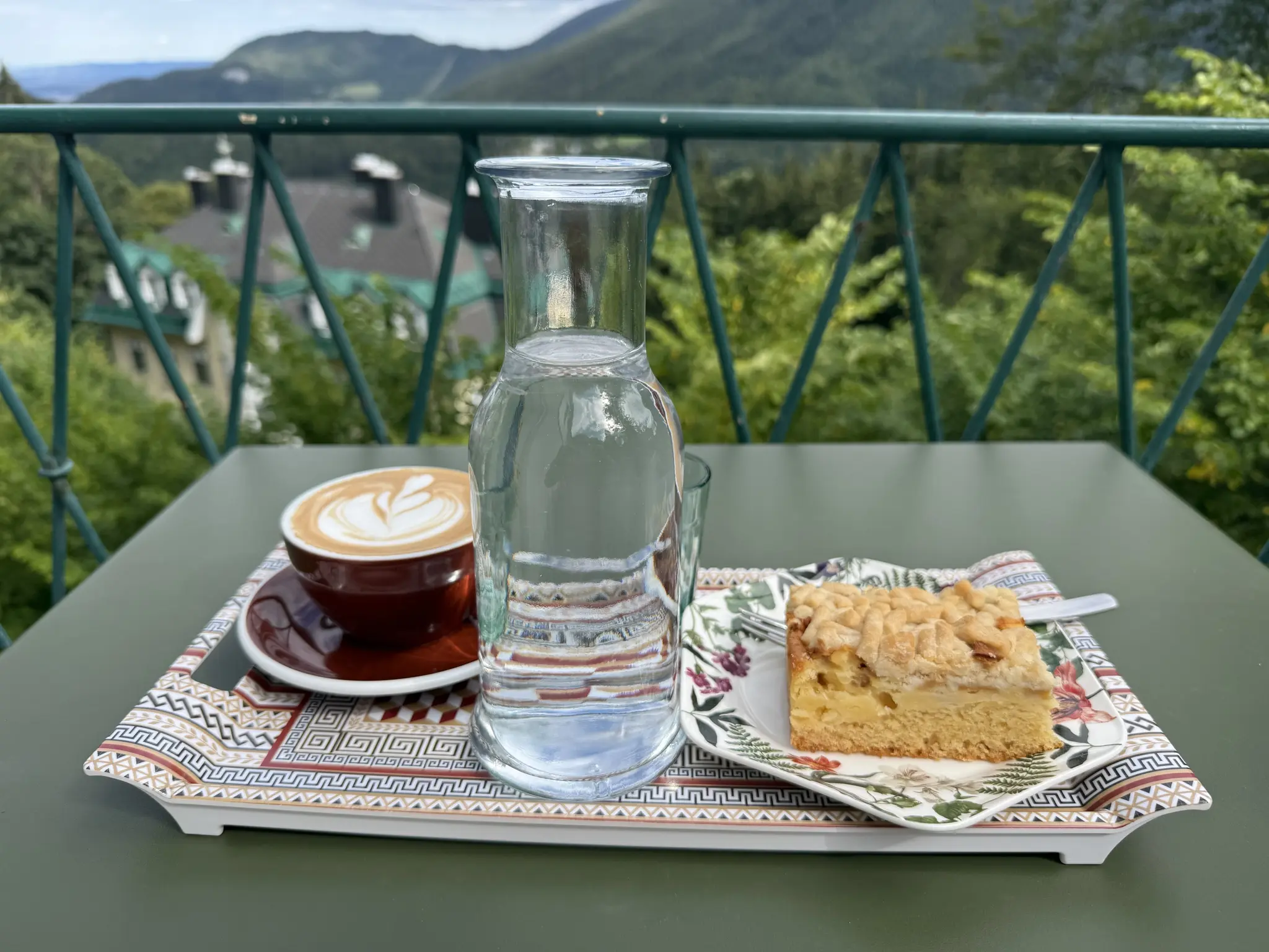 Tablett mit Kaffee, Wasser und Apfelstreuselkuchen auf einem Tisch auf Terasse mit Bergausblick
