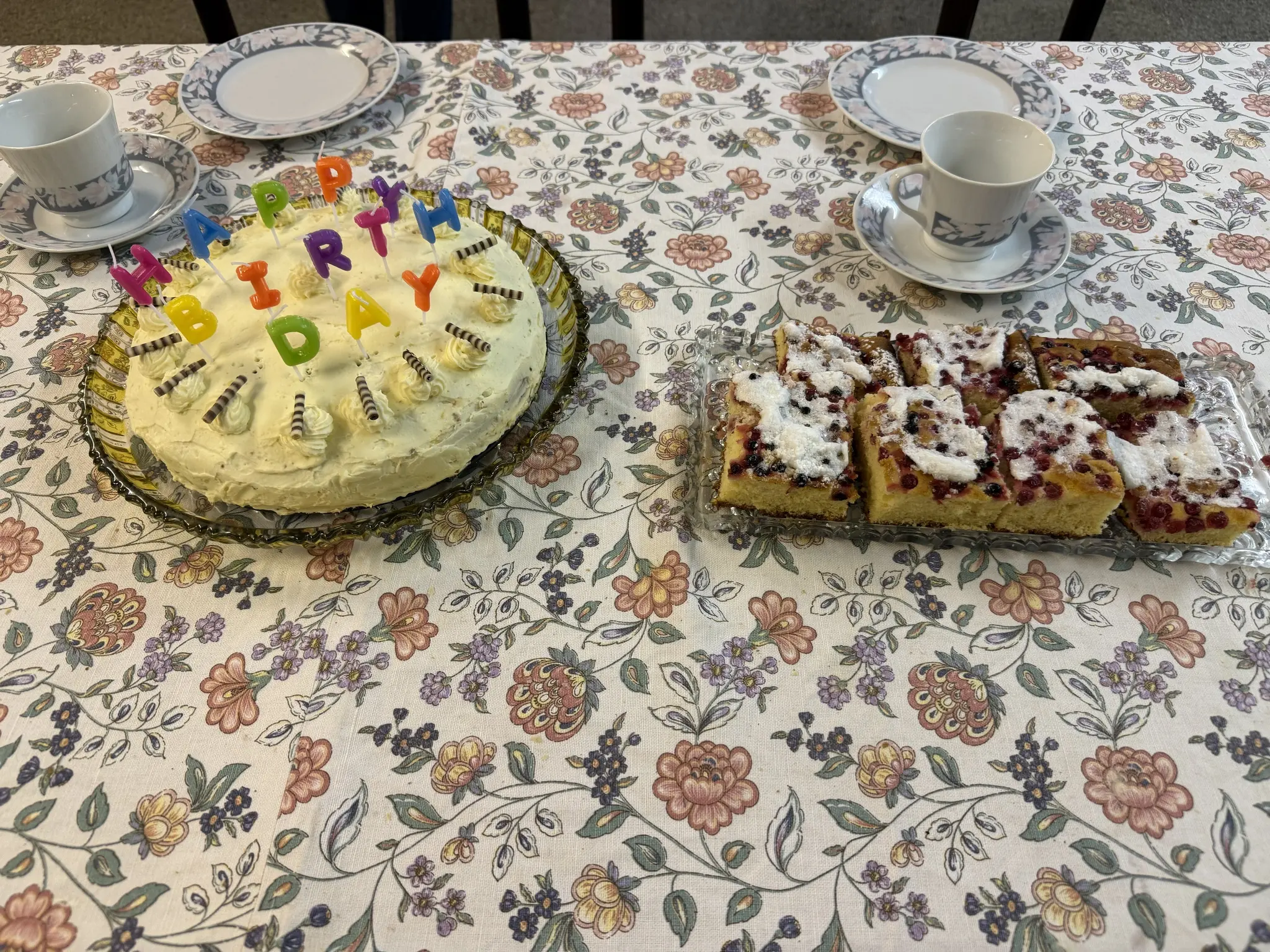 Tisch mit altlodischer Blumentischdecke, darauf eine Geburtstagstorte und ein Ribiselkuchen. Dahinter Teller mit Kaffeehäferl
