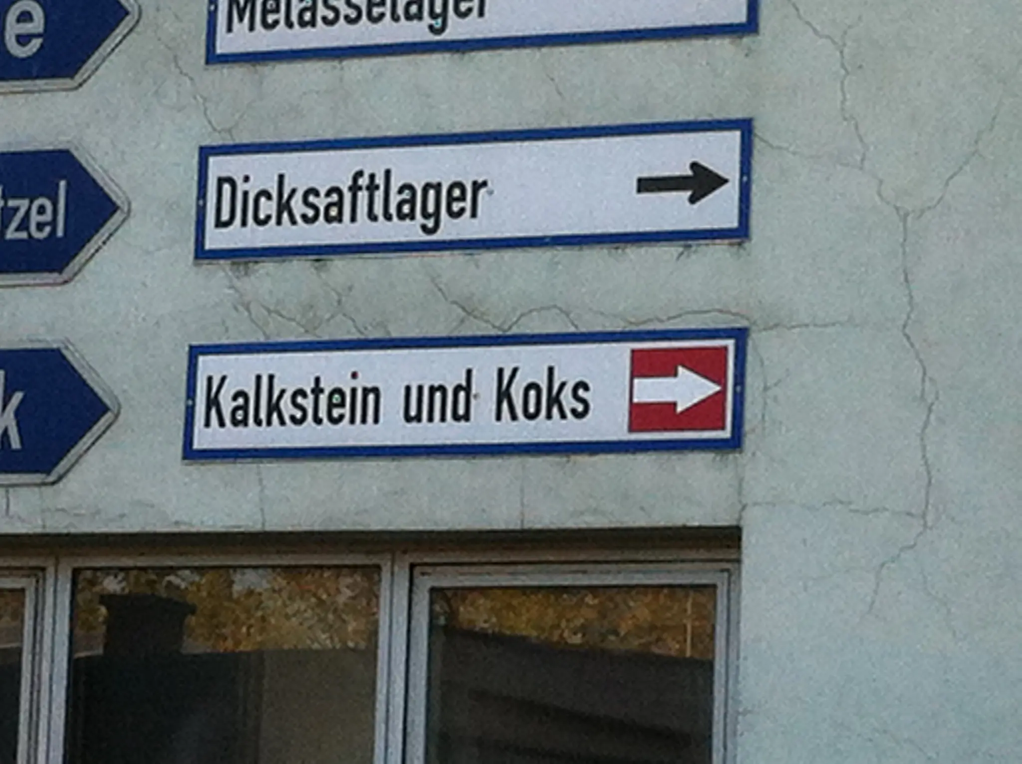 Photo of a wall featuring multiple direction signs. They are in German, and say "Melasselager", "Dicksaftlager", and "Kalkstein und Koks" respectively.