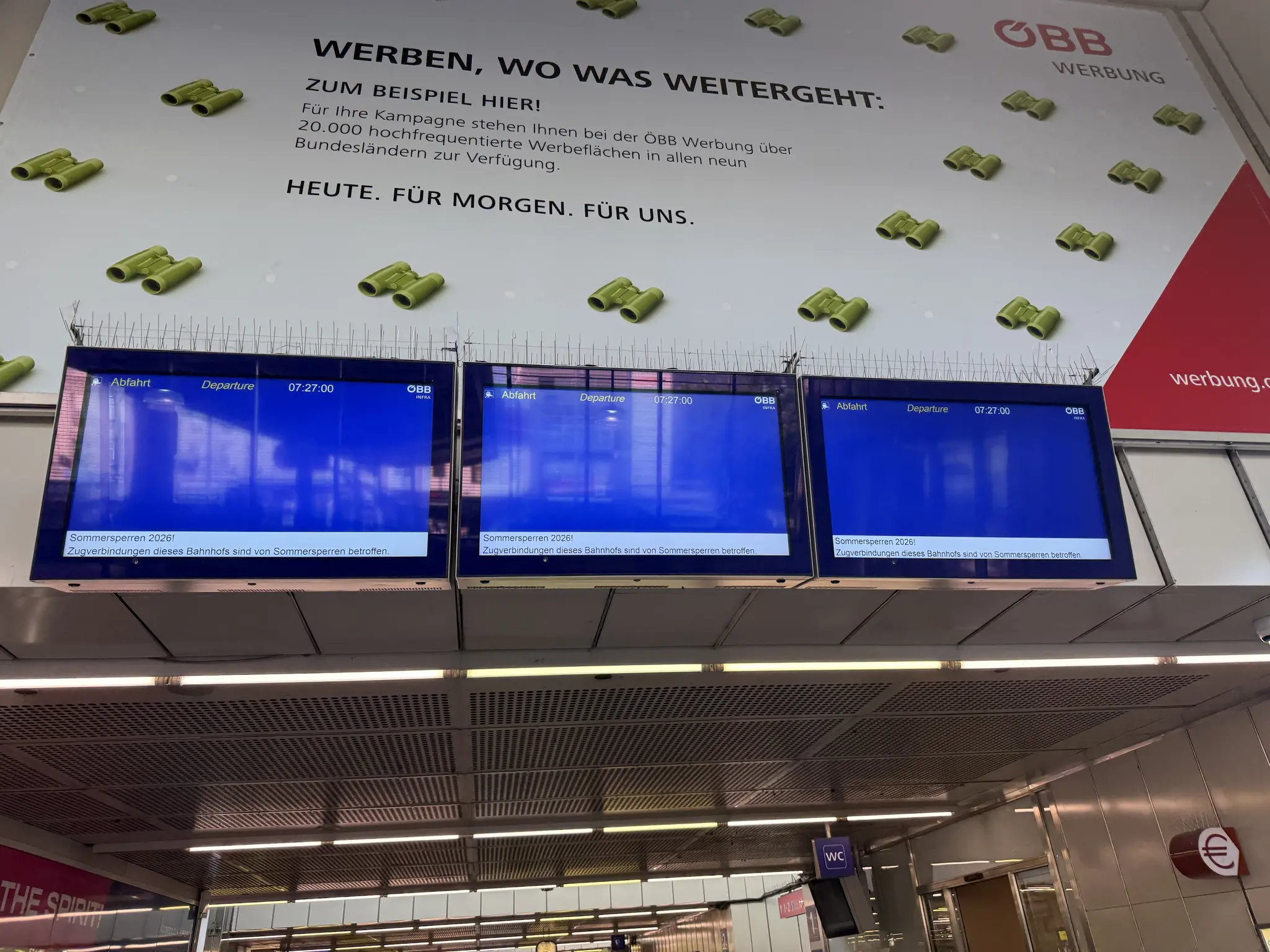 Eingangshalle vom Bahnhof Floridsdorf; drei Abfahrtsmonitore leer, sie zeigen nur oben die Uhrzeit und unten die Info zu den Sommersperren an. Darüber ein ÖBB Werbung Banner