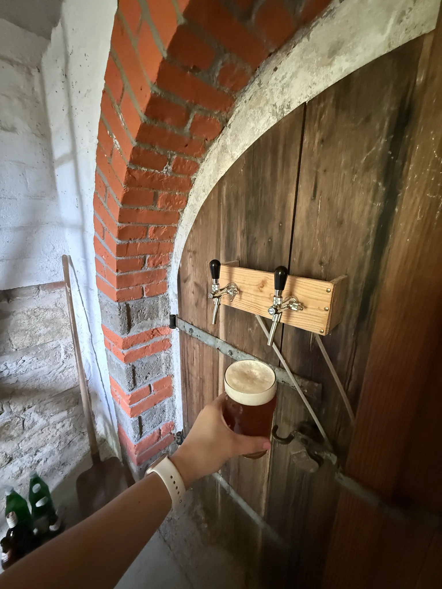 Tür von einem Weinkeller mit gemauertem Bogen. An der Tür sind die Zapfhähne fürs Bier montiert. Ein Glas rotes Bier in die Kamera gehalten