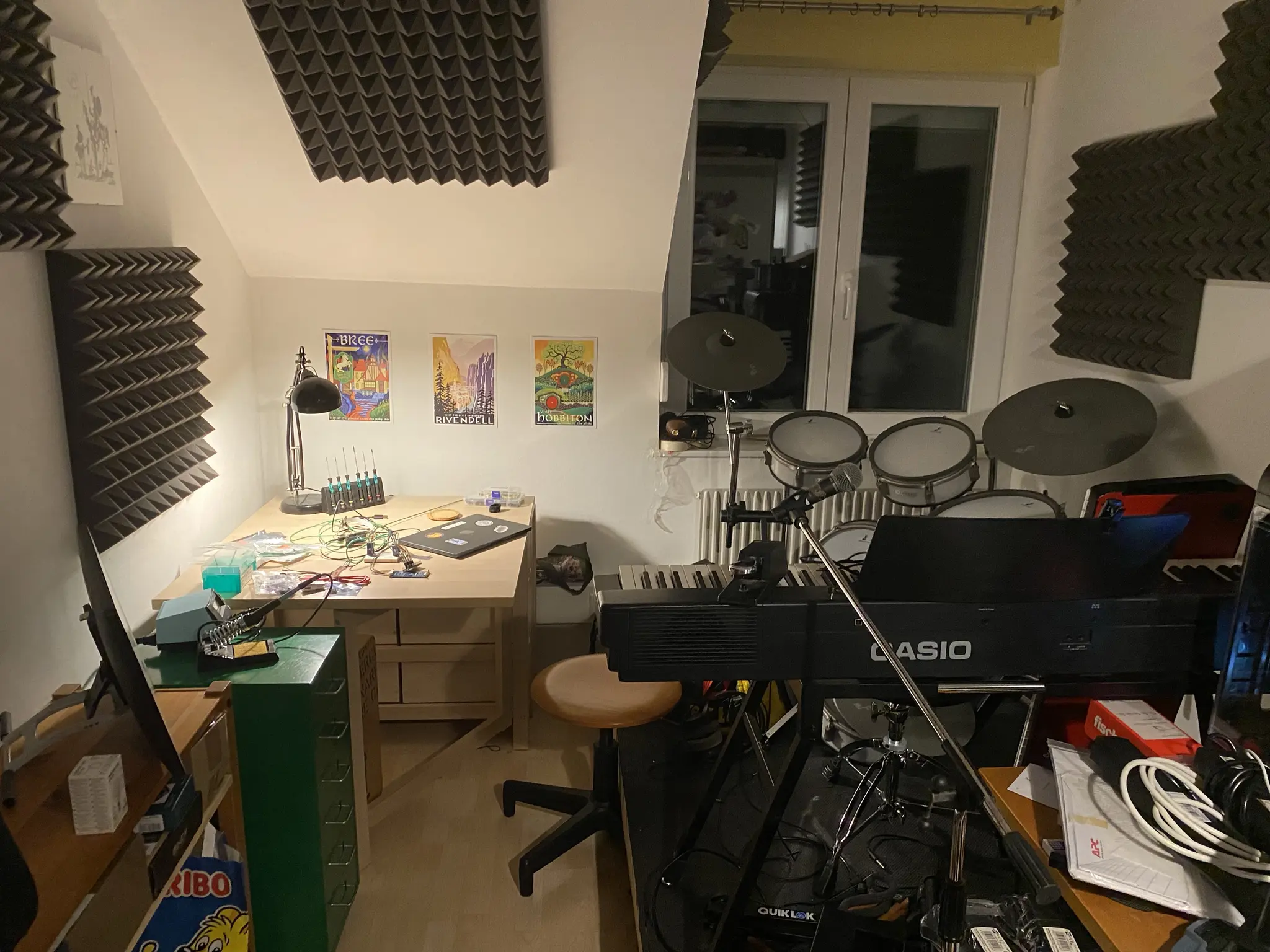 Picture of a room with a drumset to the right, and a desk with a lamp to the left. There are a bunch of electronics and cables on the table. 