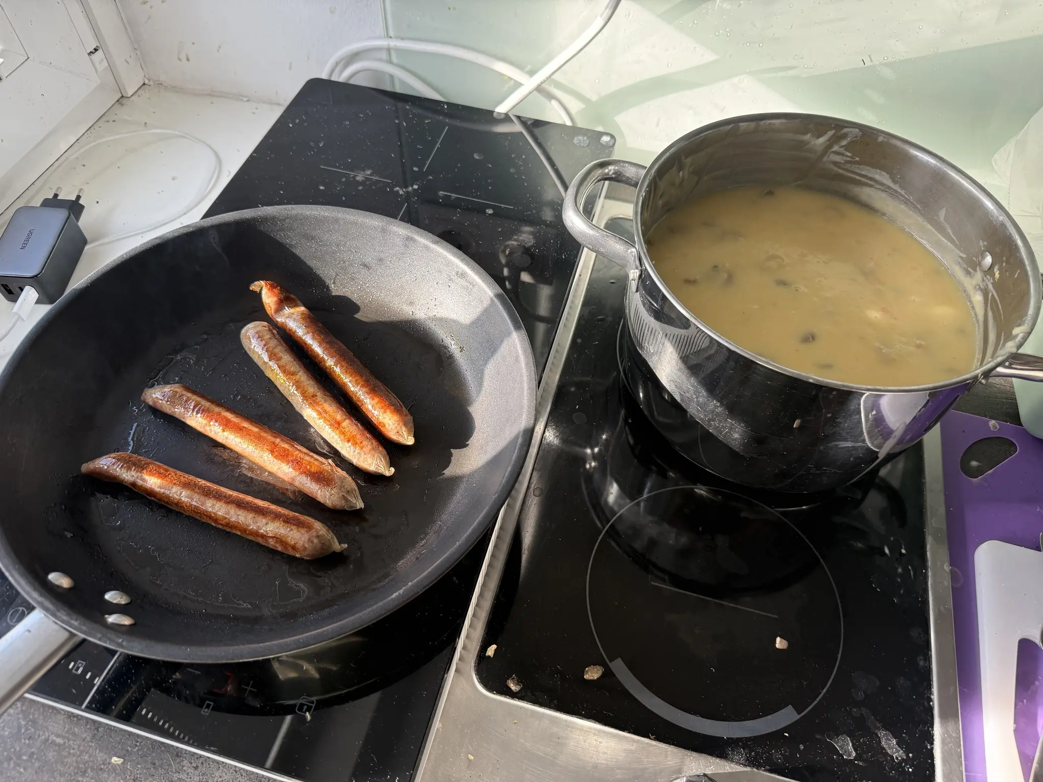 Pfanne auf einer Induktionsplatte mit veganen Bratwürstel, daneben auf einer anderen Platte ein großer Topf mit Erdäfpelsuppe