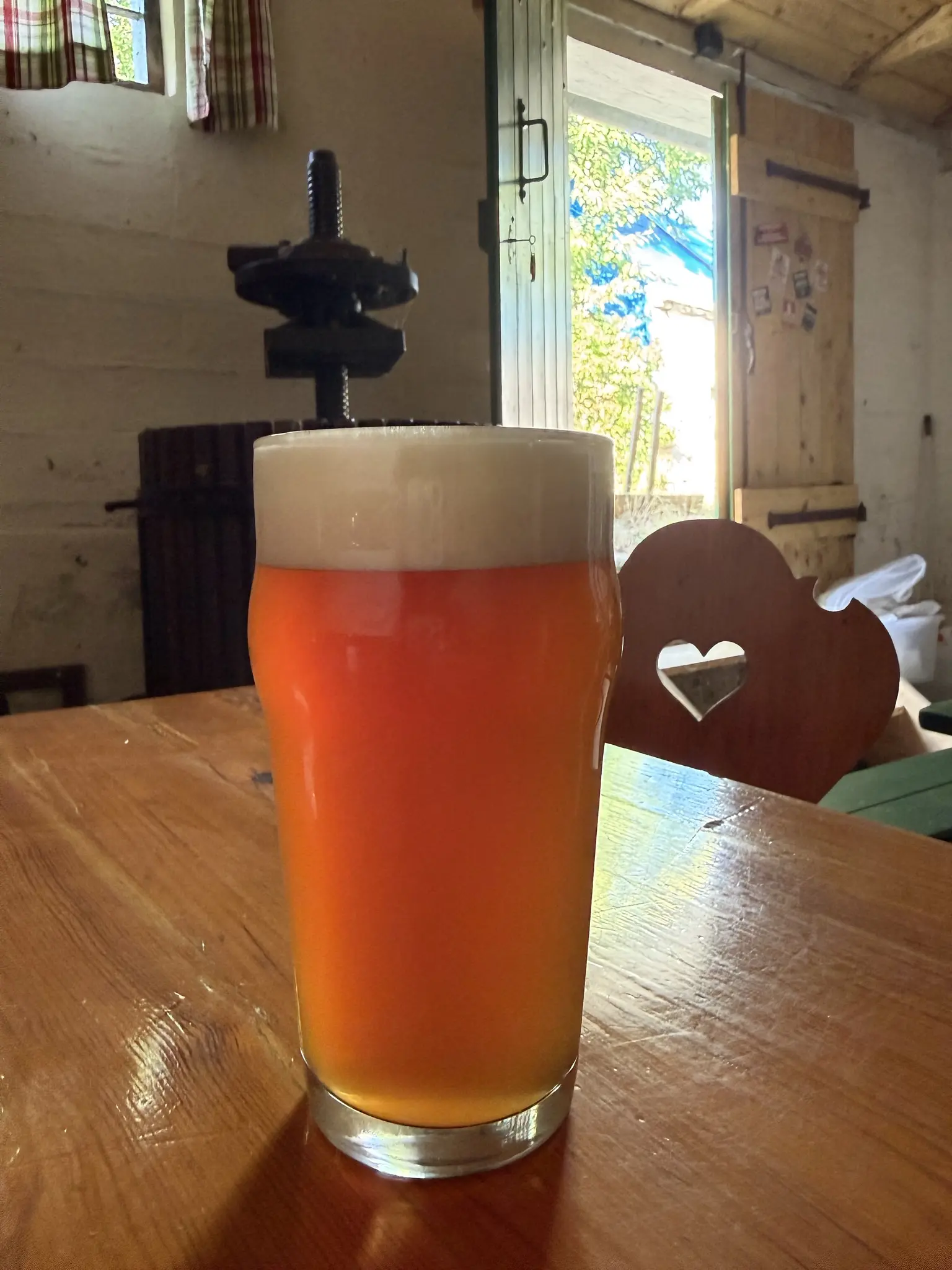 Pint-Glas mit rotem Bier mit schöner Schaumkrone auf einem Holztisch, dahinter eine kleine Weinpresse und die weiße Ziegeleand von einem Presshaus.