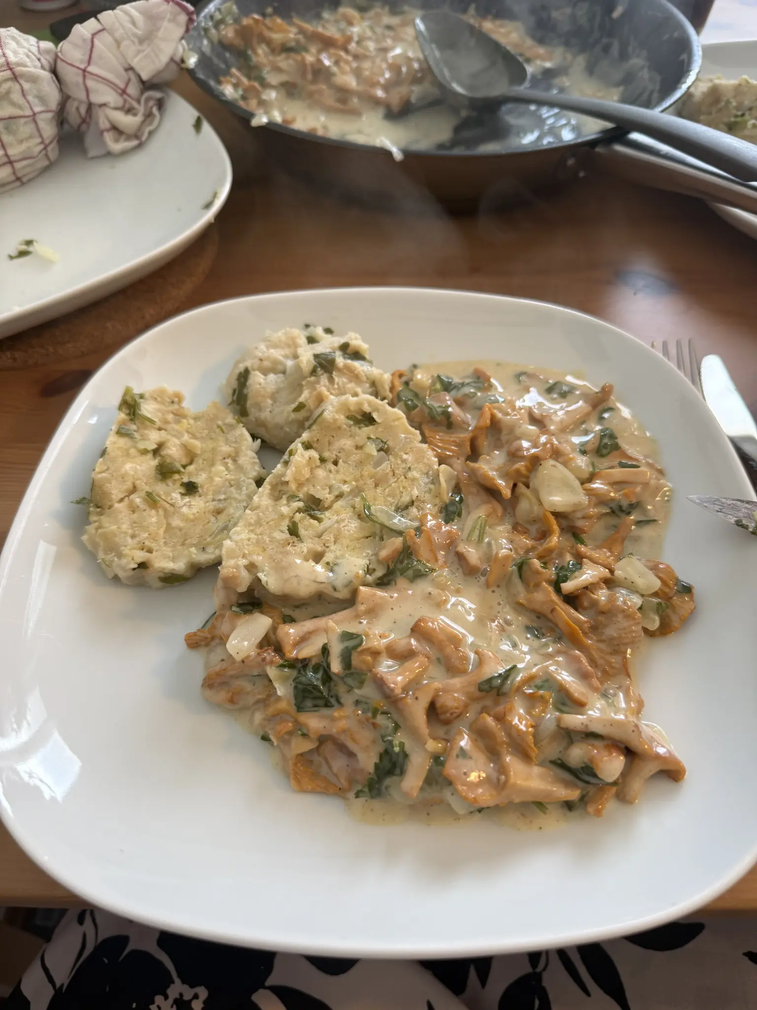 Teller mit Eierschwammerlsauce und Serviettenknödel. Dahinter eine Pfanne mit der Sauce und ein Teller, auf der noch eine zugeknotete Stoffserviette ist