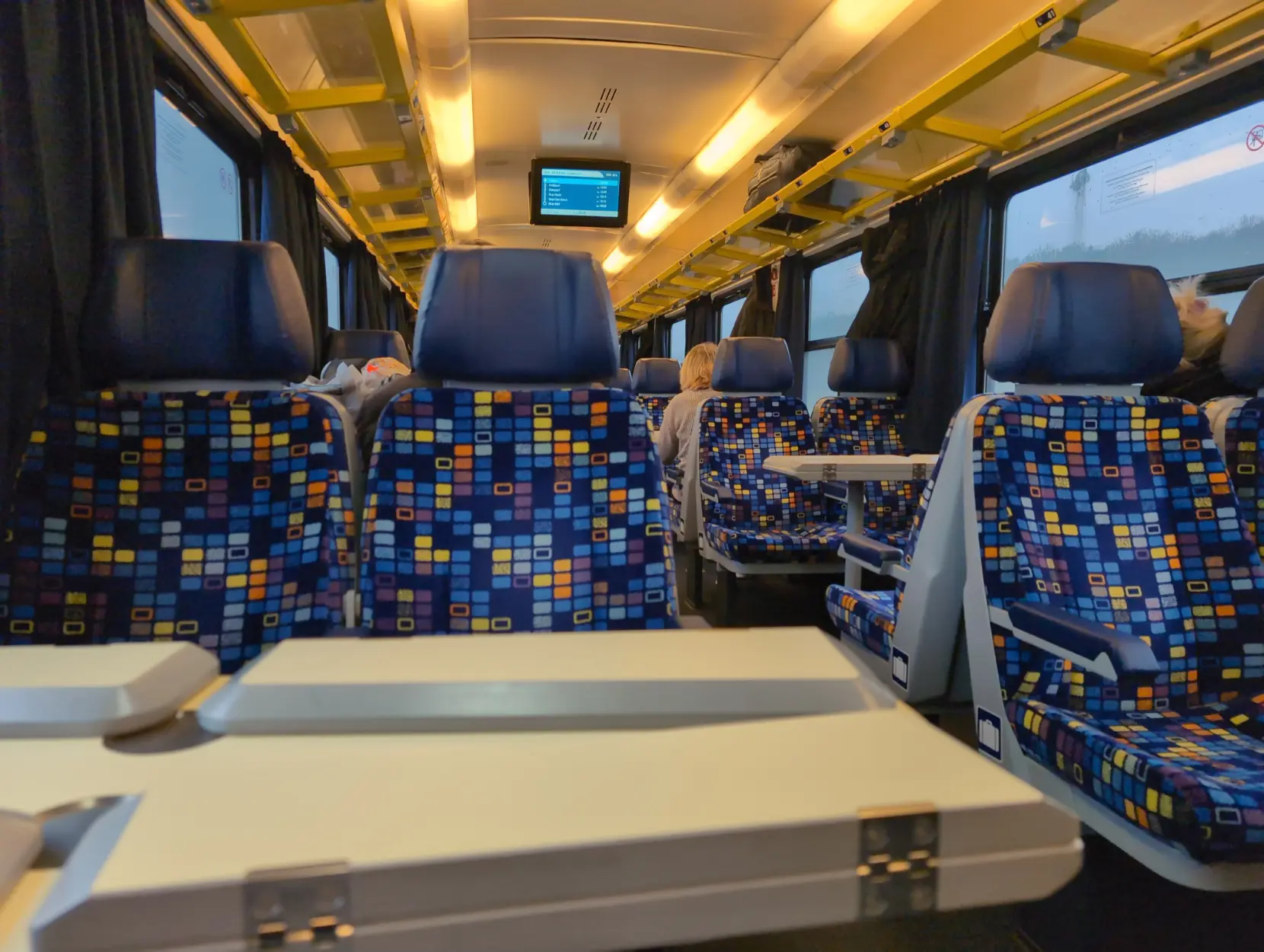 Foto vom Interieur eines MAV-Eurofima-Waggons. Die Sitze sind in 4ern angeordnet und die Überzüge sind blau mit einem Muster aus verschiedenfarbigen Rechtecken. Draußen ist schemenhaft eine Landschaft zu erkennen.