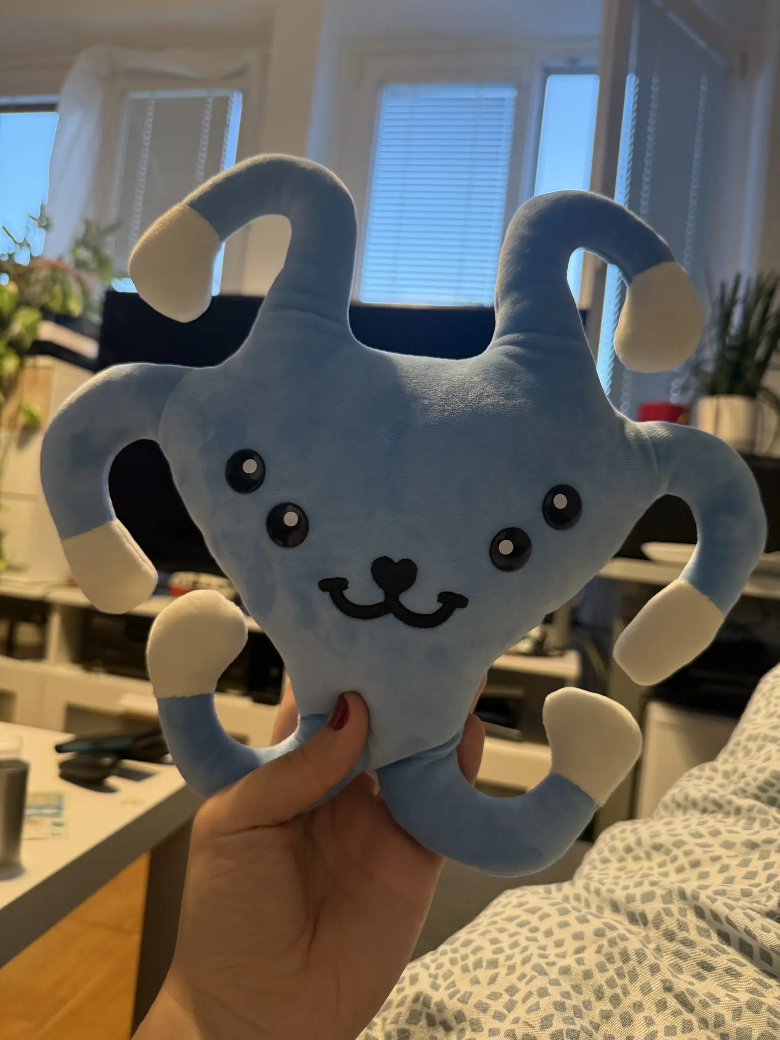 Blue alien plushie hold into the camera with 4 eyes, and are heart shaped nose with a 3-shaped mouth underneath. It‘s called Starpal from Bethesda‘s Starfield. In the background a living room with a TV